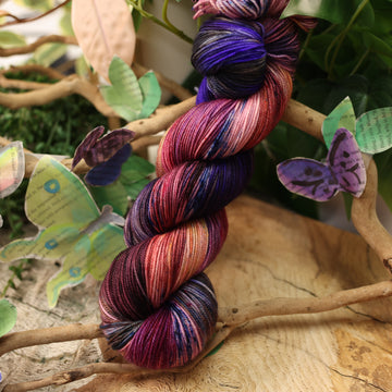 a dark purple, pink, and red colored Sport yarn with some speckles on it, on top of a plant with bare branches and leaves in the back. along with painted butterflies and leaves made of book pages, all on top of a wooden table.