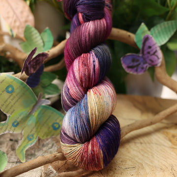a dark purple, pink, and red colored square fingering yarn with some speckles on it, on top of a plant with bare branches and leaves in the back. along with painted butterflies and leaves made of book pages, all on top of a wooden table.