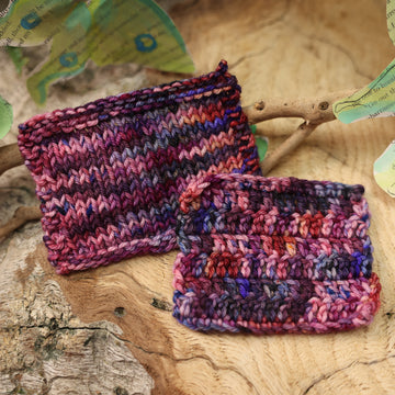 a dark purple, pink, and red colored knit and crochet swatches with some speckles on it, on top of a plant with bare branches and leaves in the back. along with painted butterflies and leaves made of book pages, all on top of a wooden table.