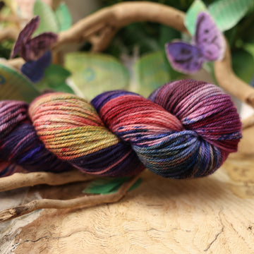 a dark purple, pink, and red colored worsted yarn with some speckles on it, on top of a plant with bare branches and leaves in the back. along with painted butterflies and leaves made of book pages, all on top of a wooden table.