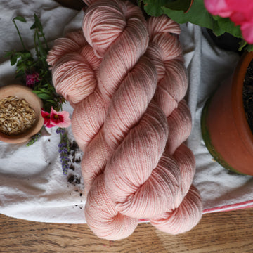 A light pink colored Dk yarn skein on top of a white tablecloth with some pink flowers and rice grains and a plant pot next to it.