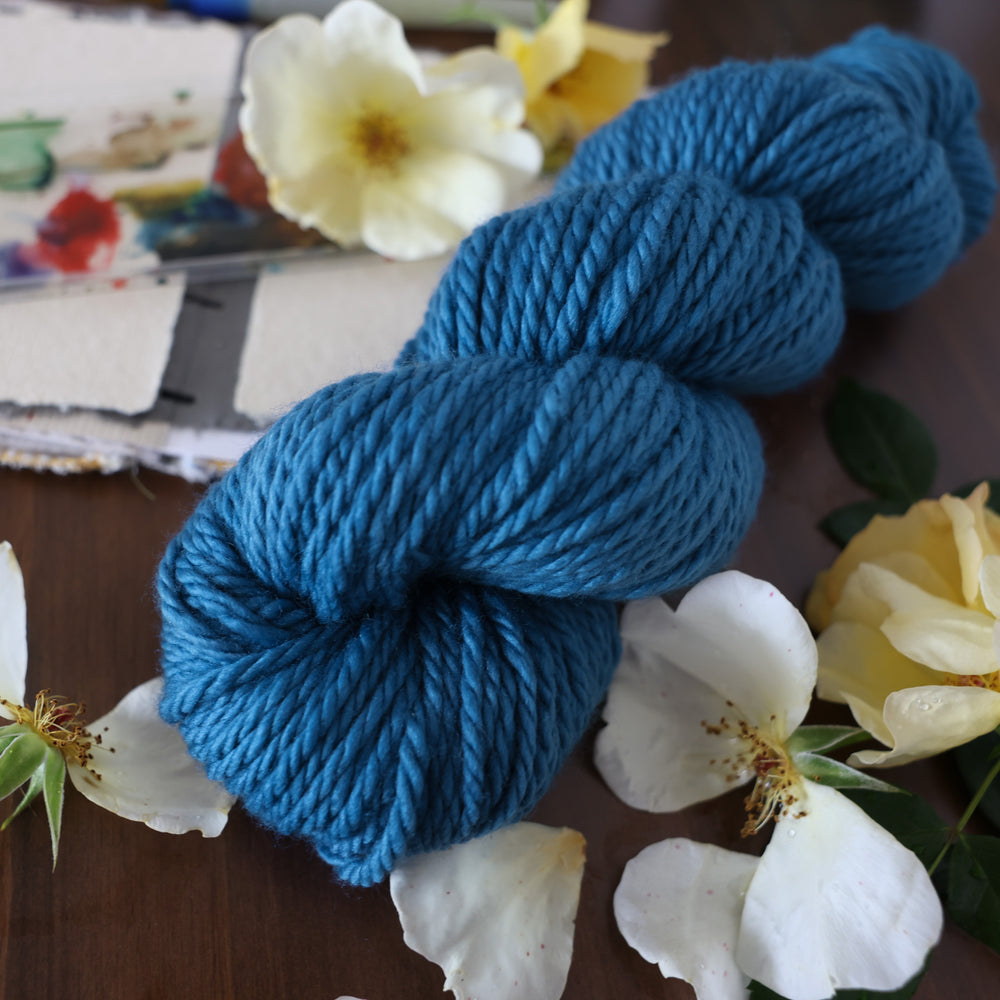 Bulky yarn colored a navy colored blue. This is on top of a brown table with a watercolor paint pallet on top of some canvas paper and some white and yellow flowers.
