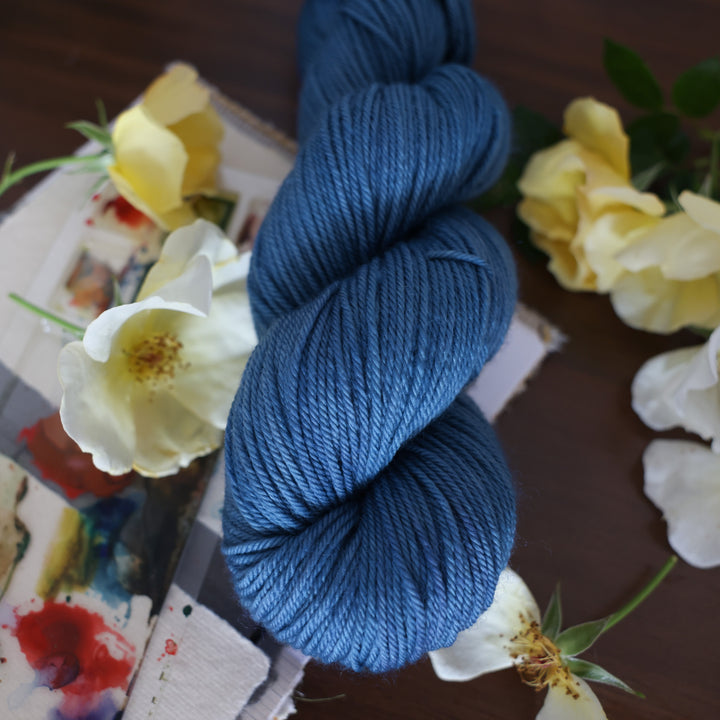 Dk yarn colored a navy colored blue. This is on top of a brown table with a watercolor paint pallet on top of some canvas paper and some white and yellow flowers.