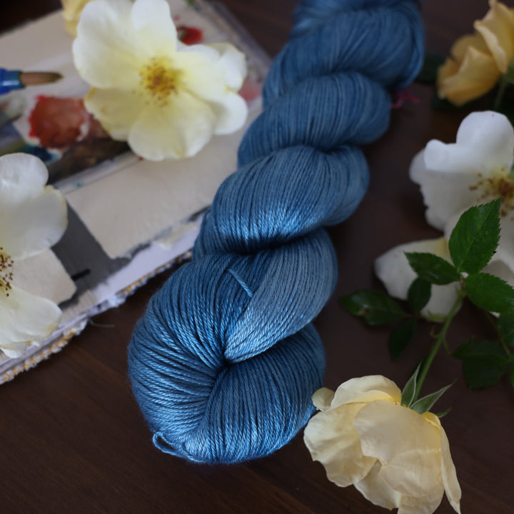 Silk yarn colored a navy colored blue. This is on top of a brown table with a watercolor paint pallet on top of some canvas paper and some white and yellow flowers.