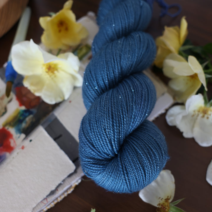 Sparkle DK yarn colored a navy colored blue. This is on top of a brown table with a watercolor paint pallet on top of some canvas paper and some white and yellow flowers.