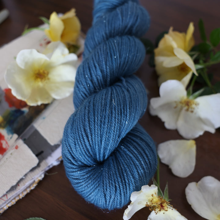 Sparkle DK yarn colored a navy colored blue. This is on top of a brown table with a watercolor paint pallet on top of some canvas paper and some white and yellow flowers.
