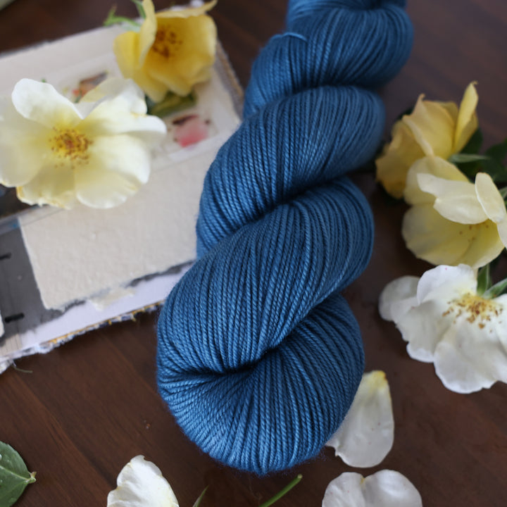 Sport yarn colored a navy colored blue. This is on top of a brown table with a watercolor paint pallet on top of some canvas paper and some white and yellow flowers.