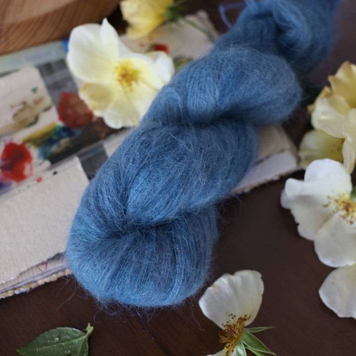 Suri yarn colored a navy colored blue. This is on top of a brown table with a watercolor paint pallet on top of some canvas paper and some white and yellow flowers.