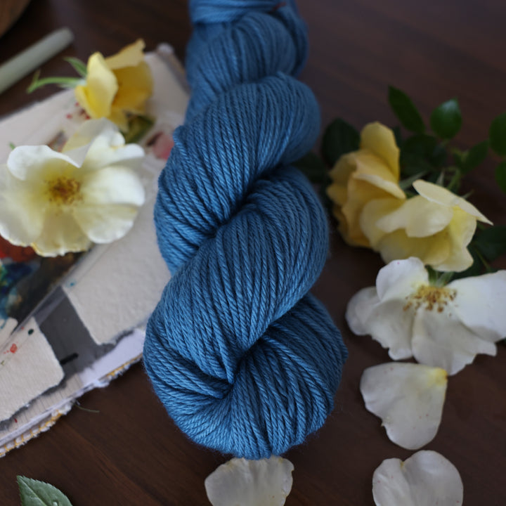 Worsted yarn colored a navy colored blue. This is on top of a brown table with a watercolor paint pallet on top of some canvas paper and some white and yellow flowers.