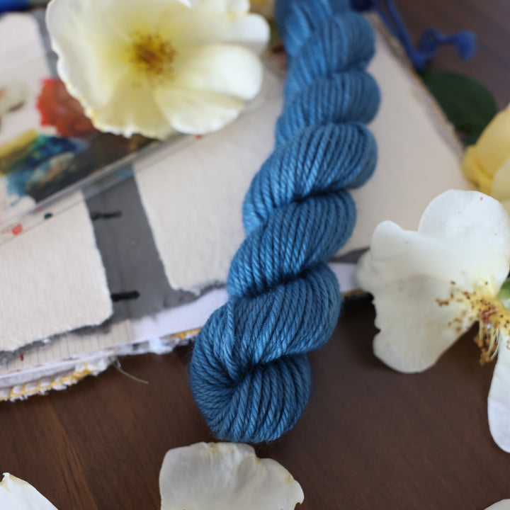 DK mini yarn colored a navy colored blue. This is on top of a brown table with a watercolor paint pallet on top of some canvas paper and some white and yellow flowers.