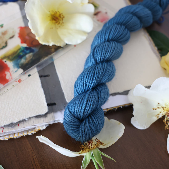 Fingering mini yarn colored a navy colored blue. This is on top of a brown table with a watercolor paint pallet on top of some canvas paper and some white and yellow flowers.