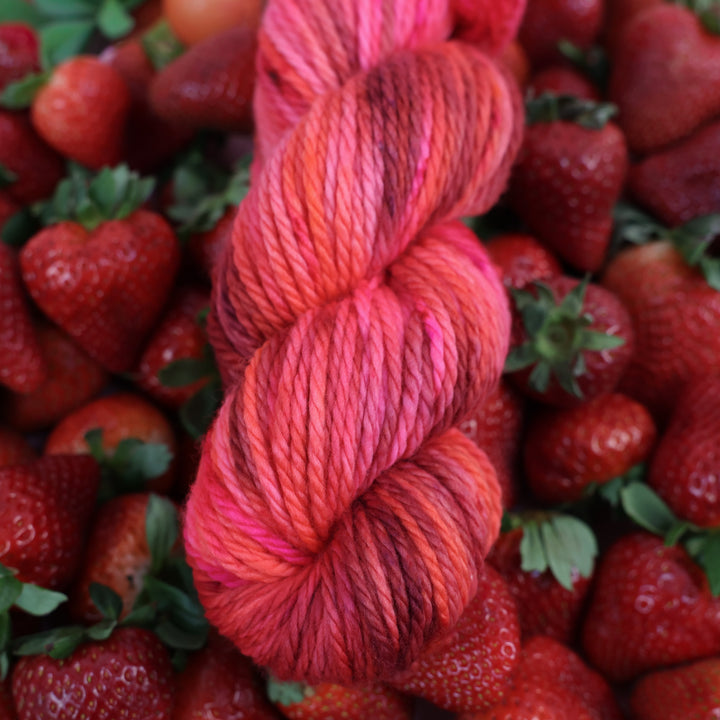 Bulky yarn that is colored with different shades of pink like bright pink, and light pink, and a dark red and dark pink. This is on top of lots of ripe strawberries.
