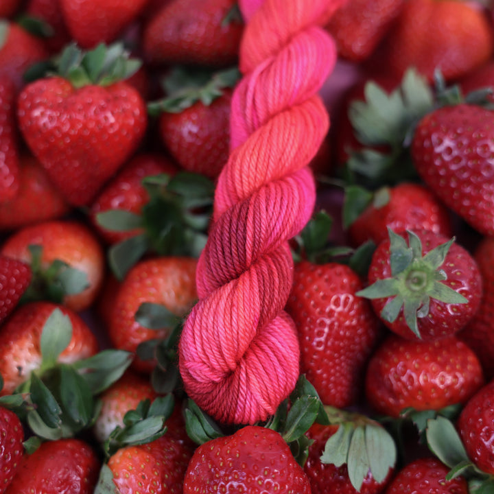 DK mini yarn that is colored with different shades of pink like bright pink, and light pink, and a dark red and dark pink. This is on top of lots of ripe strawberries.