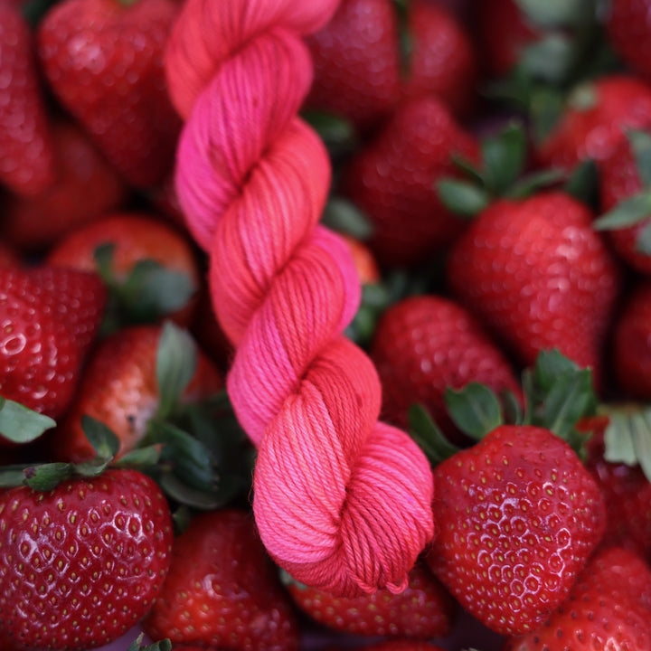 Fingering mini yarn that is colored with different shades of pink like bright pink, and light pink, and a dark red and dark pink. This is on top of lots of ripe strawberries.