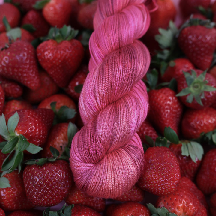 Silk yarn that is colored with different shades of pink like bright pink, and light pink, and a dark red and dark pink. This is on top of lots of ripe strawberries.