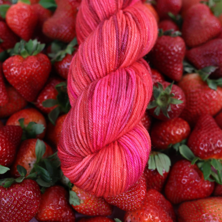 Sparkle DK yarn that is colored with different shades of pink like bright pink, and light pink, and a dark red and dark pink. This is on top of lots of ripe strawberries.