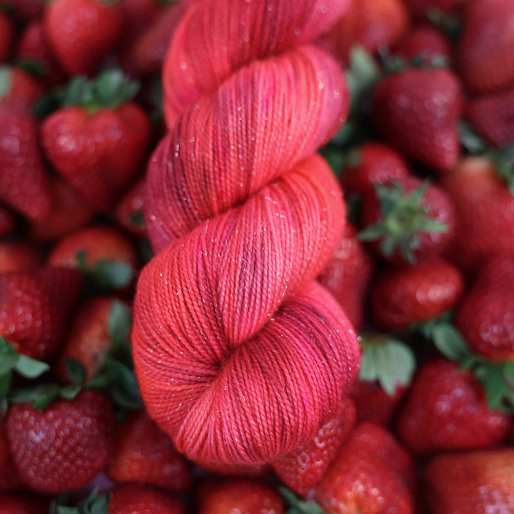 Sparkle fingering yarn that is colored with different shades of pink like bright pink, and light pink, and a dark red and dark pink. This is on top of lots of ripe strawberries.