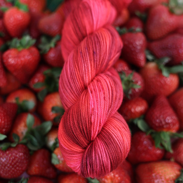 Sport yarn that is colored with different shades of pink like bright pink, and light pink, and a dark red and dark pink. This is on top of lots of ripe strawberries.
