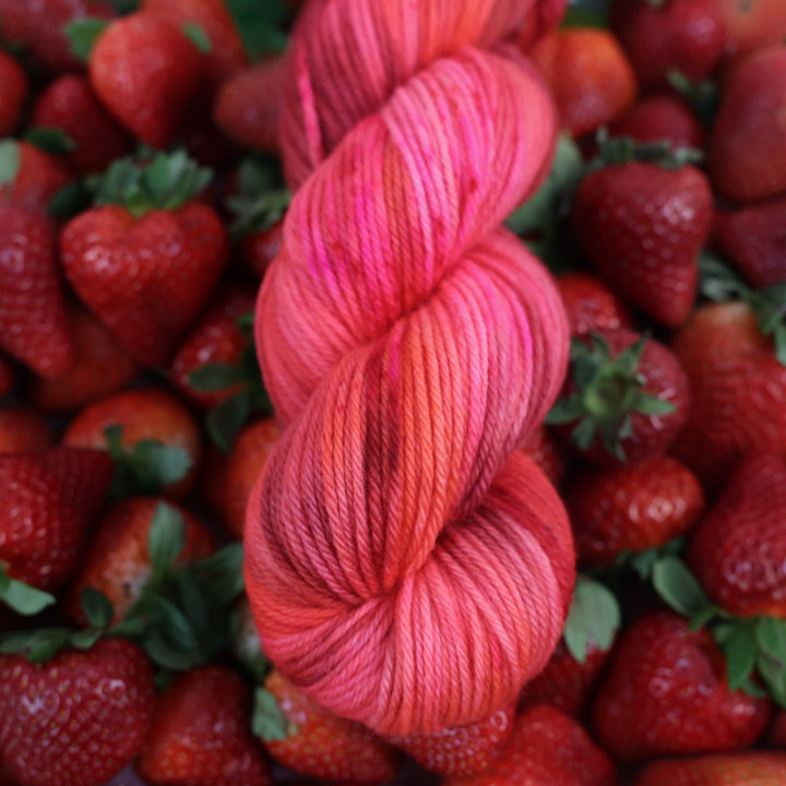 Worsted yarn that is colored with different shades of pink like bright pink, and light pink, and a dark red and dark pink. This is on top of lots of ripe strawberries.