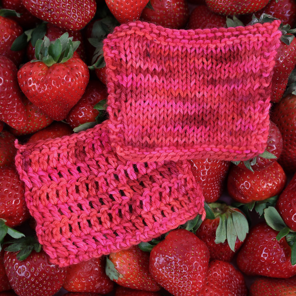 Crochet and knit swatches that are colored with different shades of pink like bright pink, and light pink, and a dark red and dark pink. This is on top of lots of ripe strawberries.
