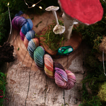 S-mini yarn colored with a hot pink, light blue, dark blue/purple, with some green and orange/brown colors, on a wooden table with some moss surrounding it and a wooden circle with mushrooms on it and a small glass beetle.