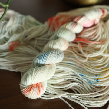 S-mini white yarn with mixed colors of light pink purple and red/orange and brown with speckles, on top of an unwound version of that same yarn. all on top of a dark wooden table with a couple of leaves of a plant overhead in a gold pot.