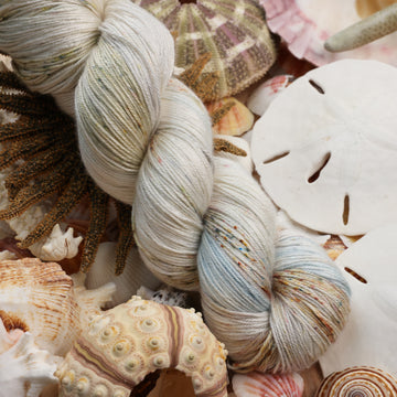 Silk yarn that is colored a very light blue and cream and has different colored dark brown and dark blue speckles. This yarn is on top of a bunch of different types  of light colored shells and sea stars and sand dollars.