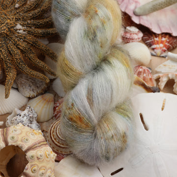 Suri yarn that is colored a very light blue and cream and has different colored dark brown and dark blue speckles. This yarn is on top of a bunch of different types  of light colored shells and sea stars and sand dollars.