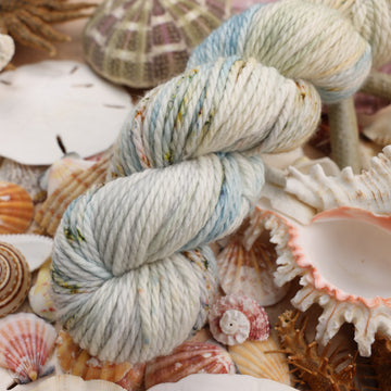 Bulky yarn that is colored a very light blue and cream and has different colored dark brown and dark blue speckles. This yarn is on top of a bunch of different types  of light colored shells and sea stars and sand dollars.