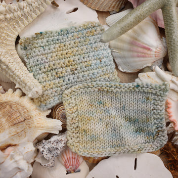 Knit and crochet swatches that are colored a very light blue and cream and has different colored dark brown and dark blue speckles. These swatches are on top of a bunch of different types  of light colored shells and sea stars and sand dollars.