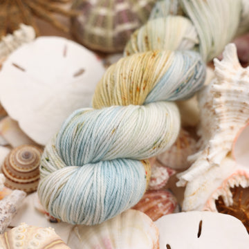 DK yarn that is colored a very light blue and cream and has different colored dark brown and dark blue speckles. This yarn is on top of a bunch of different types  of light colored shells and sea stars and sand dollars.