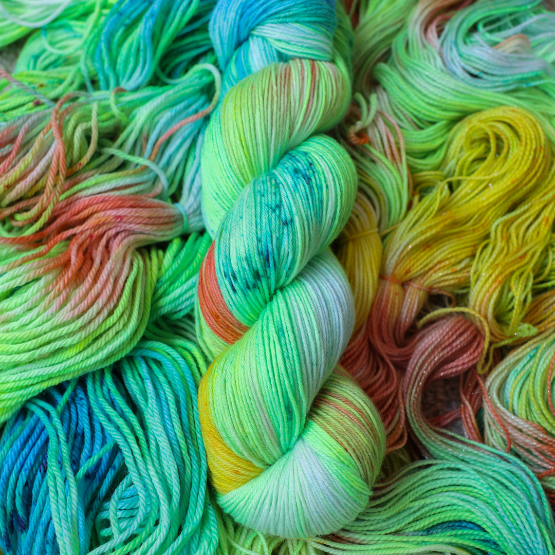 Sock yarn colored different colors like a light neon green, light blue, some streaks of white, some red, and some yellow. This skein is on top of unwound skeins of the same color way.