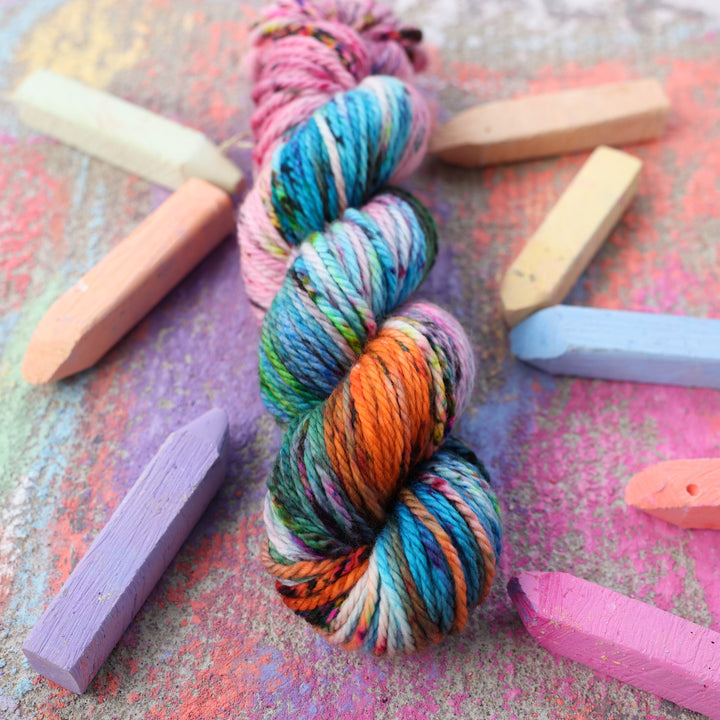 Bulky yarn colored with colors like light and dark pink, a salmon orange and red, green, purple, different shades of blue, and speckles of all these colors. this skein is on a sidewalk with different colored drawings underneath and sticks of sidewalk chalk next to them.