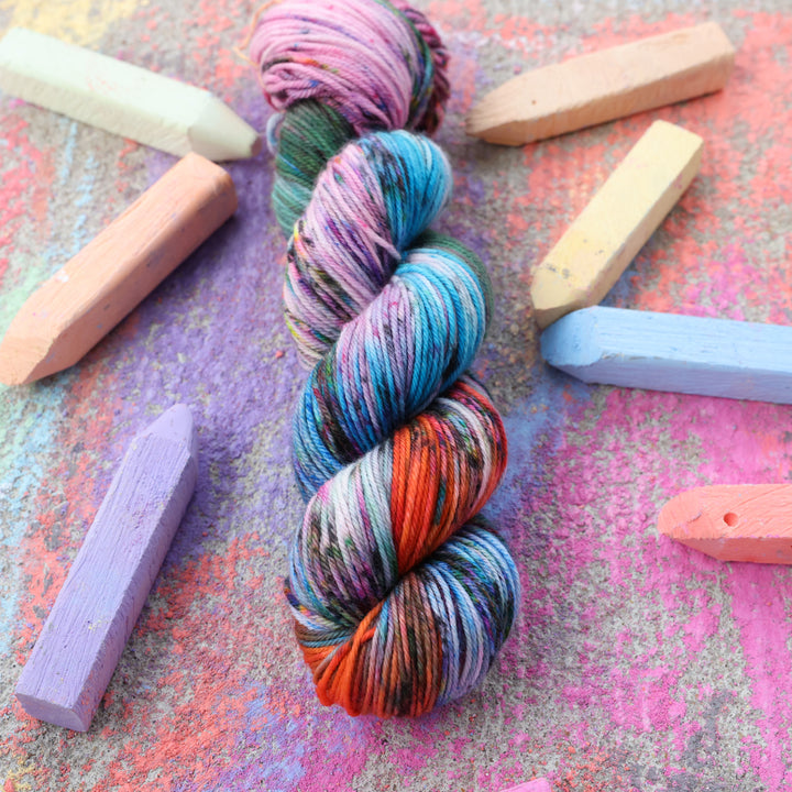 DK yarn colored with colors like light and dark pink, a salmon orange and red, green, purple, different shades of blue, and speckles of all these colors. this skein is on a sidewalk with different colored drawings underneath and sticks of sidewalk chalk next to them.