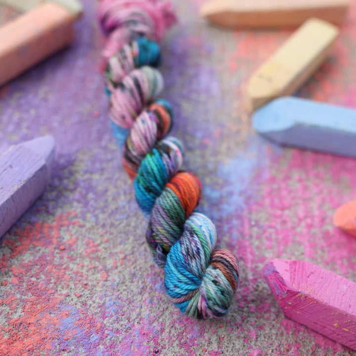 DK mini yarn colored with colors like light and dark pink, a salmon orange and red, green, purple, different shades of blue, and speckles of all these colors. this skein is on a sidewalk with different colored drawings underneath and sticks of sidewalk chalk next to them.