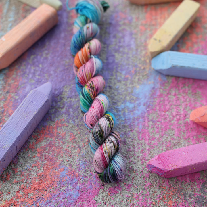 Fingering mini yarn colored with colors like light and dark pink, a salmon orange and red, green, purple, different shades of blue, and speckles of all these colors. this skein is on a sidewalk with different colored drawings underneath and sticks of sidewalk chalk next to them.