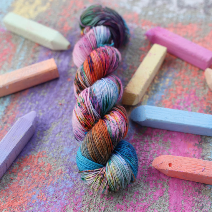 Fingering yarn colored with colors like light and dark pink, a salmon orange and red, green, purple, different shades of blue, and speckles of all these colors. this skein is on a sidewalk with different colored drawings underneath and sticks of sidewalk chalk next to them.