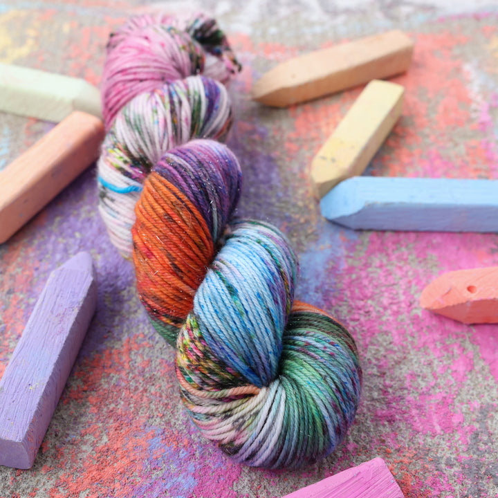 Sparkle Dk yarn colored with colors like light and dark pink, a salmon orange and red, green, purple, different shades of blue, and speckles of all these colors. this skein is on a sidewalk with different colored drawings underneath and sticks of sidewalk chalk next to them.