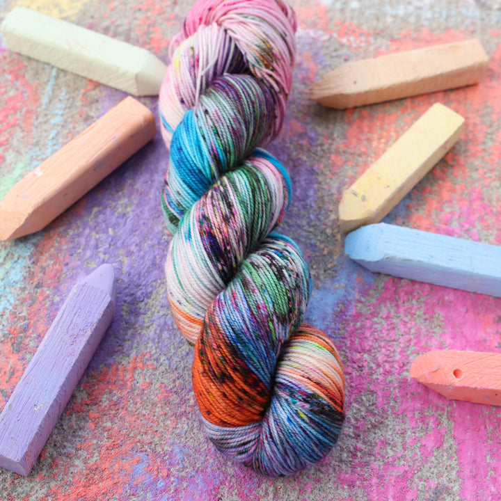 Sparkle yarn colored with colors like light and dark pink, a salmon orange and red, green, purple, different shades of blue, and speckles of all these colors. this skein is on a sidewalk with different colored drawings underneath and sticks of sidewalk chalk next to them.