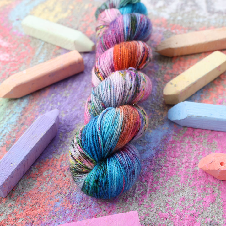 Sparkle fingering yarn colored with colors like light and dark pink, a salmon orange and red, green, purple, different shades of blue, and speckles of all these colors. this skein is on a sidewalk with different colored drawings underneath and sticks of sidewalk chalk next to them.