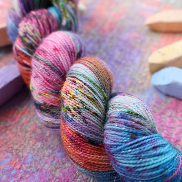 an up-close photo of sparkle fingering yarn colored with colors like light and dark pink, a salmon orange and red, green, purple, different shades of blue, and speckles of all these colors. this skein is on a sidewalk with different colored drawings underneath and sticks of sidewalk chalk next to them.