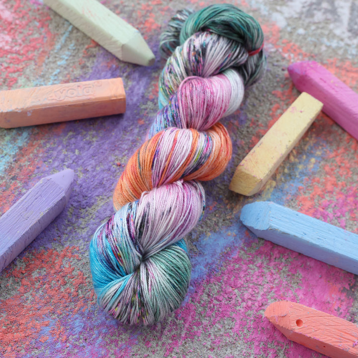 Silk yarn colored with colors like light and dark pink, a salmon orange and red, green, purple, different shades of blue, and speckles of all these colors. this skein is on a sidewalk with different colored drawings underneath and sticks of sidewalk chalk next to them.