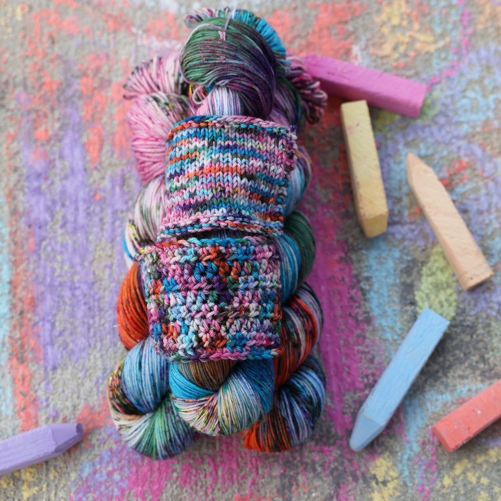 yarn and crochet and knit swatches next to each other colored with colors like light and dark pink, a salmon orange and red, green, purple, different shades of blue, and speckles of all these colors. These skeins and swatches are on a sidewalk with different colored drawings underneath and sticks of sidewalk chalk next to them.