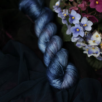 A dark blue silk yarn that has hues of grey and purple. This skein is on top of a dark purple tablecloth and dark blackish blue tablecloth with blue and purplish pink flowers laying next to the skein.