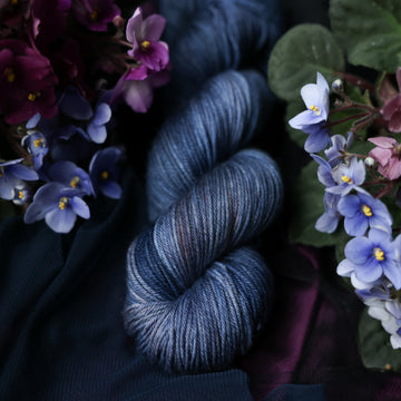 A dark and light grayish blue DK yarn that has hues of grey. This skein is on top of a dark purple tablecloth and dark blackish blue tablecloth with blue and purplish pink flowers laying next to the skein on both sides.