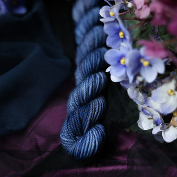 A dark blue DK mini yarn that has hues of grey and purple. This skein is on top of a dark purple tablecloth and dark blackish blue tablecloth with blue and purplish pink flowers laying next to the skein.