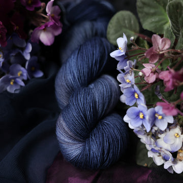 A dark blue Fingering yarn that has hues of grey. This skein is on top of a dark purple tablecloth and dark blackish blue tablecloth with blue and purplish pink flowers laying next to the skein on both sides.