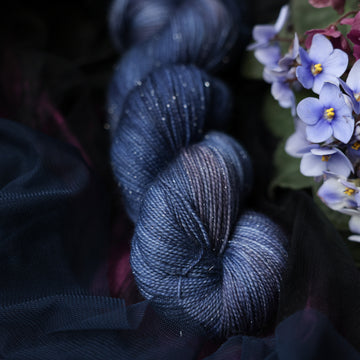 A dark blue Sparkle fingering yarn that has hues of grey. This skein is on top of a dark purple tablecloth and dark blackish blue tablecloth with blue and purplish pink flowers laying next to the skein on both sides.