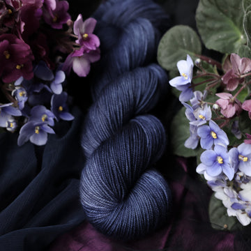 A dark blue sport yarn that has hues of grey and purple. This skein is on top of a dark purple tablecloth and dark blackish blue tablecloth with blue and purplish pink flowers laying next to the skein on both sides.