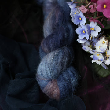 A dark blue suri yarn that has hues of grey and purple and light grayish pink. This skein is on top of a dark purple tablecloth and dark blackish blue tablecloth with blue and purplish pink flowers laying next to the skein.